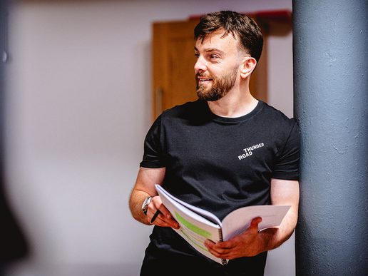 Alex Moran stands casually in a rehearsal space, leaning against a pillar while holding a marked-up script and pen. He is wearing a black "Thunder Road" t-shirt and looking to the side with a relaxed, focused expression.