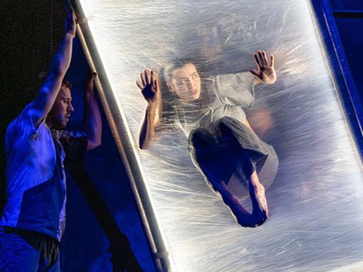 A contemporary circus performer is suspended mid-air inside a transparent vertical frame, appearing to press against a taut, elastic surface. Another performer stabilises the structure on the side, with dramatic blue lighting enhancing the surreal and immersive stage atmosphere.