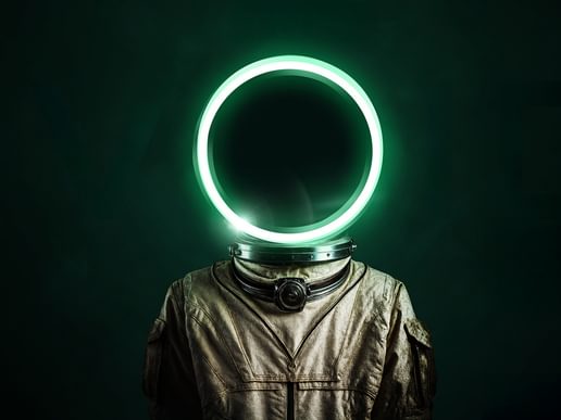 A stylised image of a figure wearing a metallic, retro-style space suit with a large glowing neon circle in place of a head. The circle emits a green light and contrasts against the dark green background, creating an otherworldly and futuristic atmosphere. The figure’s body language is neutral, and the lack of facial features adds a sense of mystery and abstraction. Likely promotional artwork for a science fiction or conceptual performance titled The Void.
